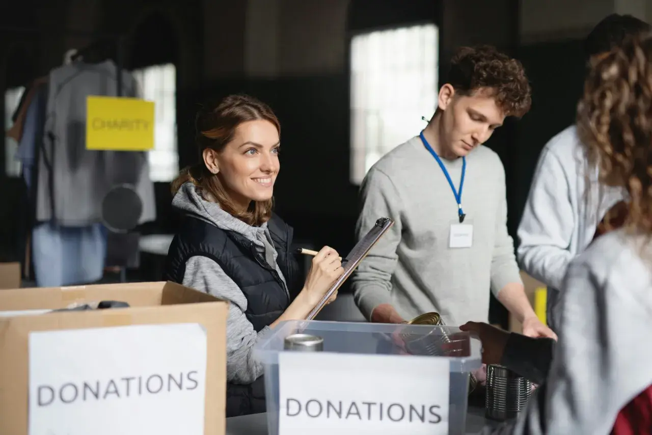 Volunteers organize donations for a free online community service. A woman smiles while checking items on a clipboard, while others sort canned goods.