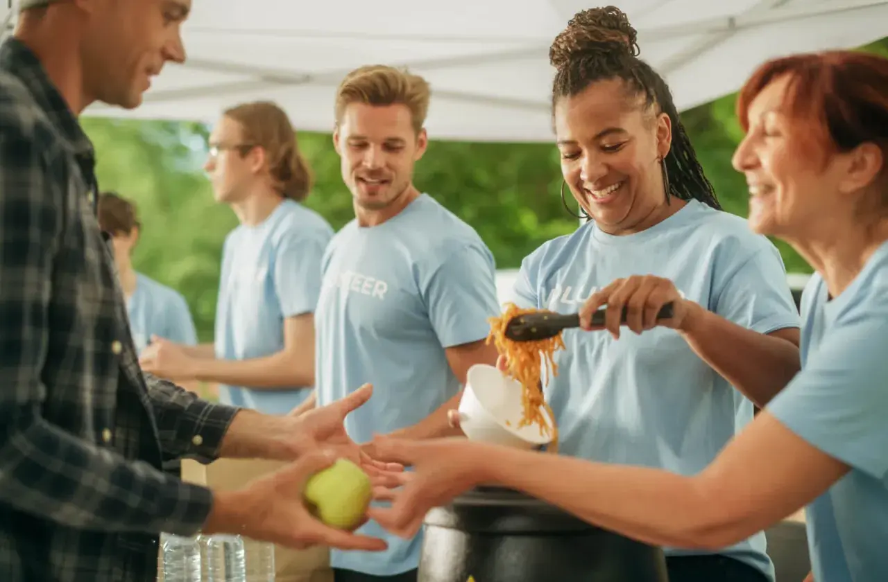 Volunteers serve food, showing what are some community services. A man receives an apple, while others get noodles.