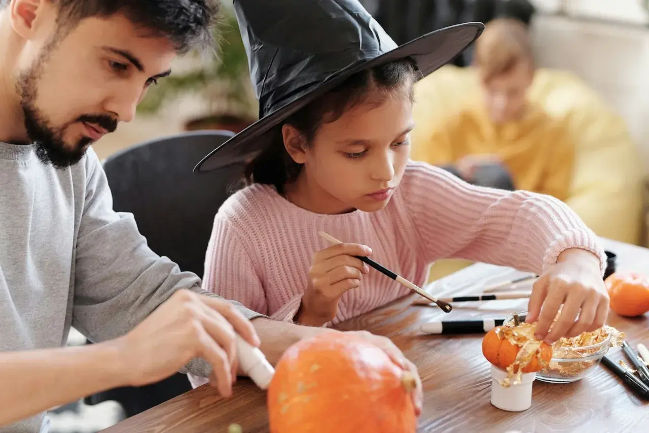 A girl in a witch hat and a man decorate pumpkins, showcasing fun Halloween fundraiser ideas.
