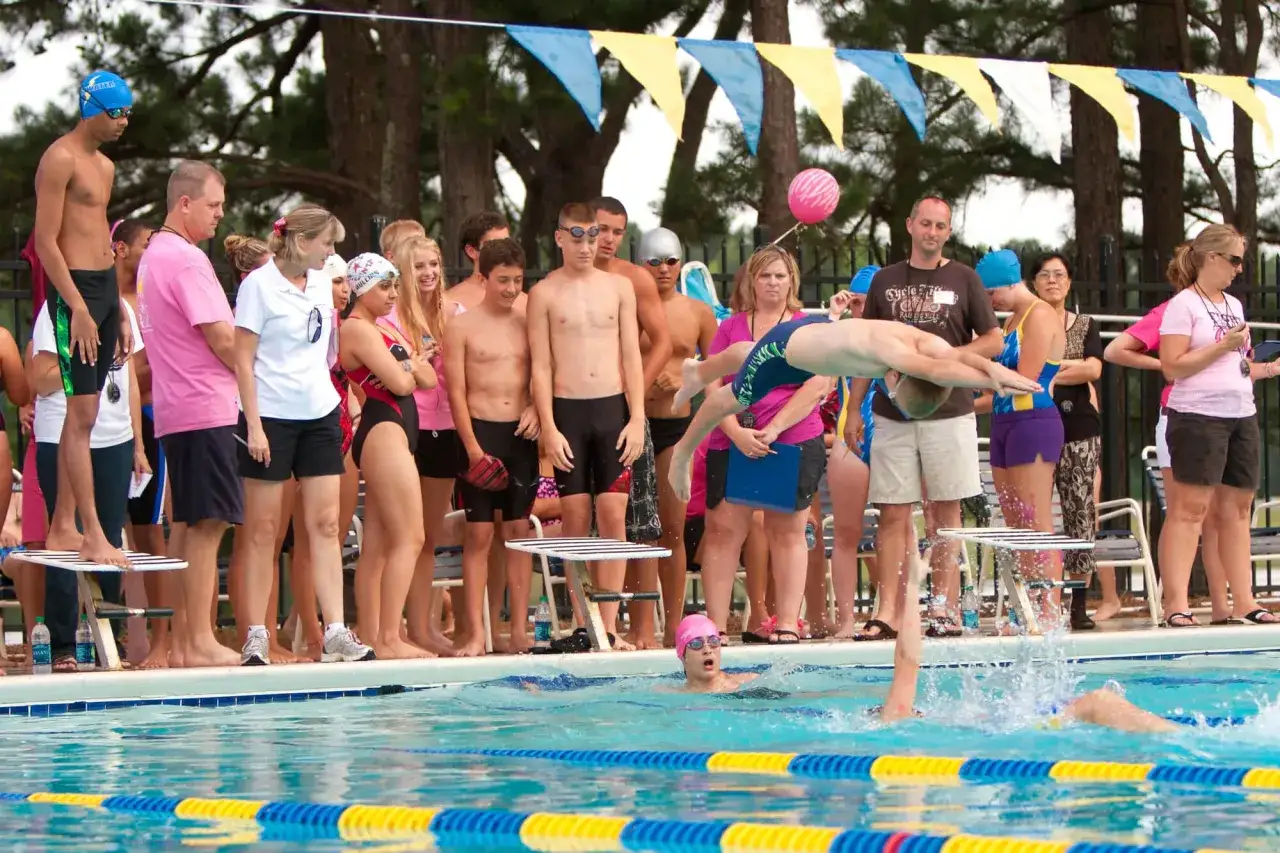 Swimmers and spectators gather for swim team fundraisers. A young swimmer dives into the pool while others watch from the deck.