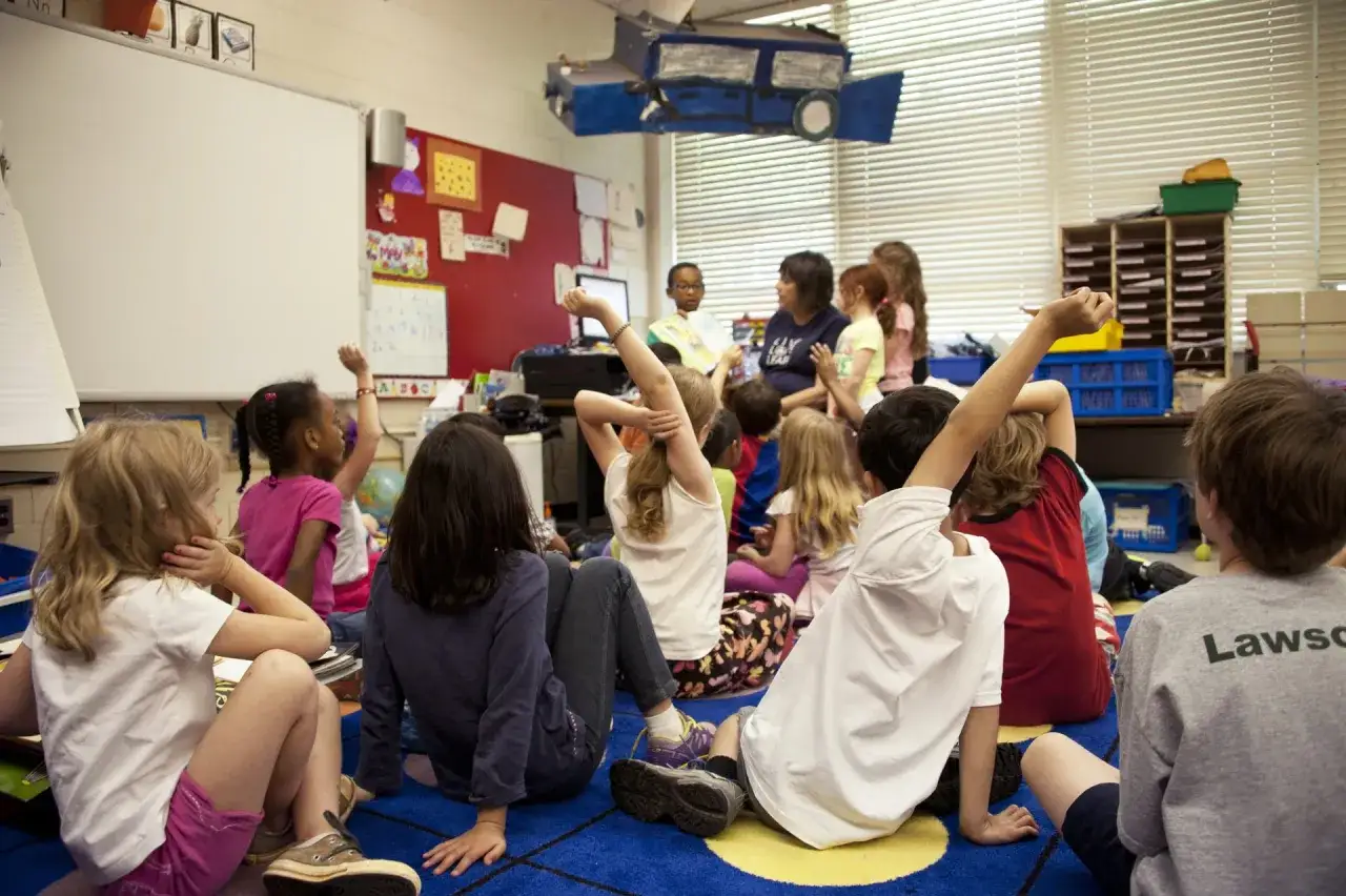 Children in a classroom, raising hands, perhaps brainstorming fun preschool fundraising ideas for new classroom supplies.