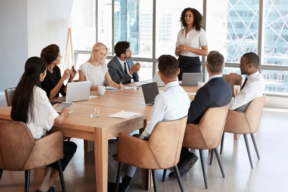 A board chair leads a meeting, guiding discussion and ensuring everyone contributes. Diverse professionals collaborate around a table.