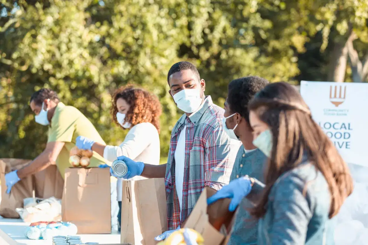 Volunteers pack food bags, showcasing business philanthropy examples. They wear masks and gloves, distributing canned goods and produce for a local food bank.
