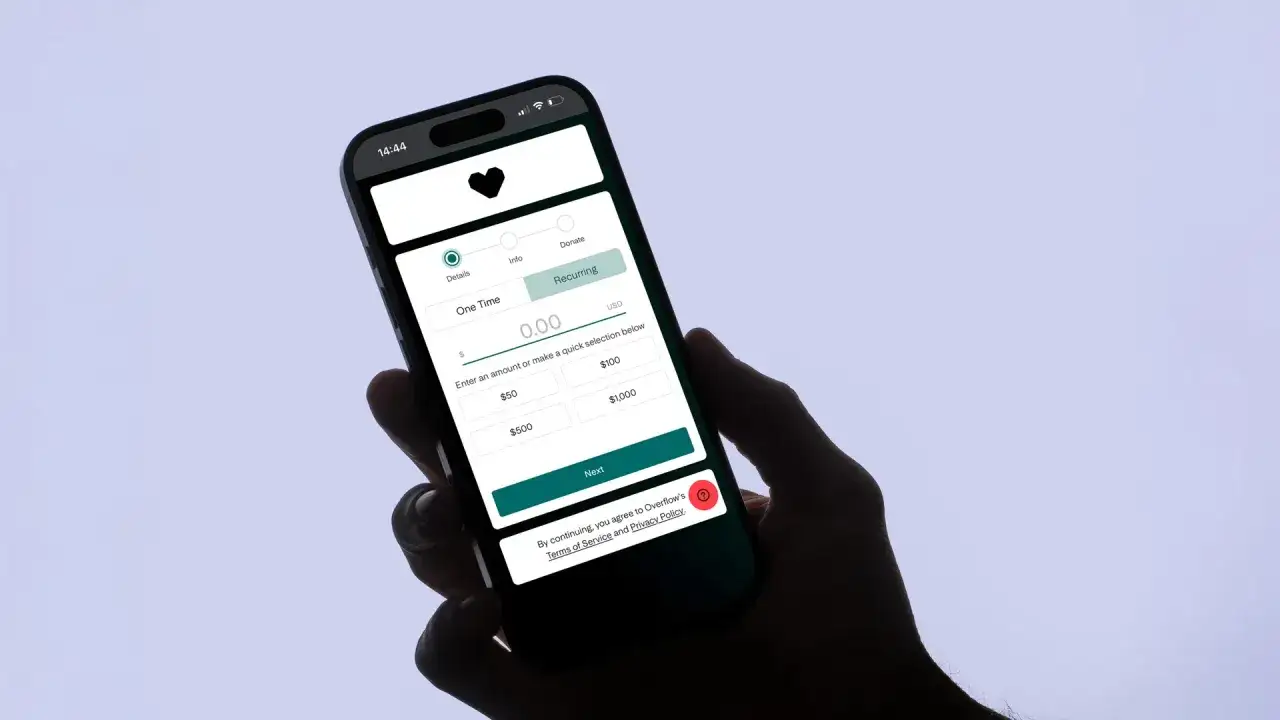 A hand holds a smartphone displaying a donation app interface. Users can select 