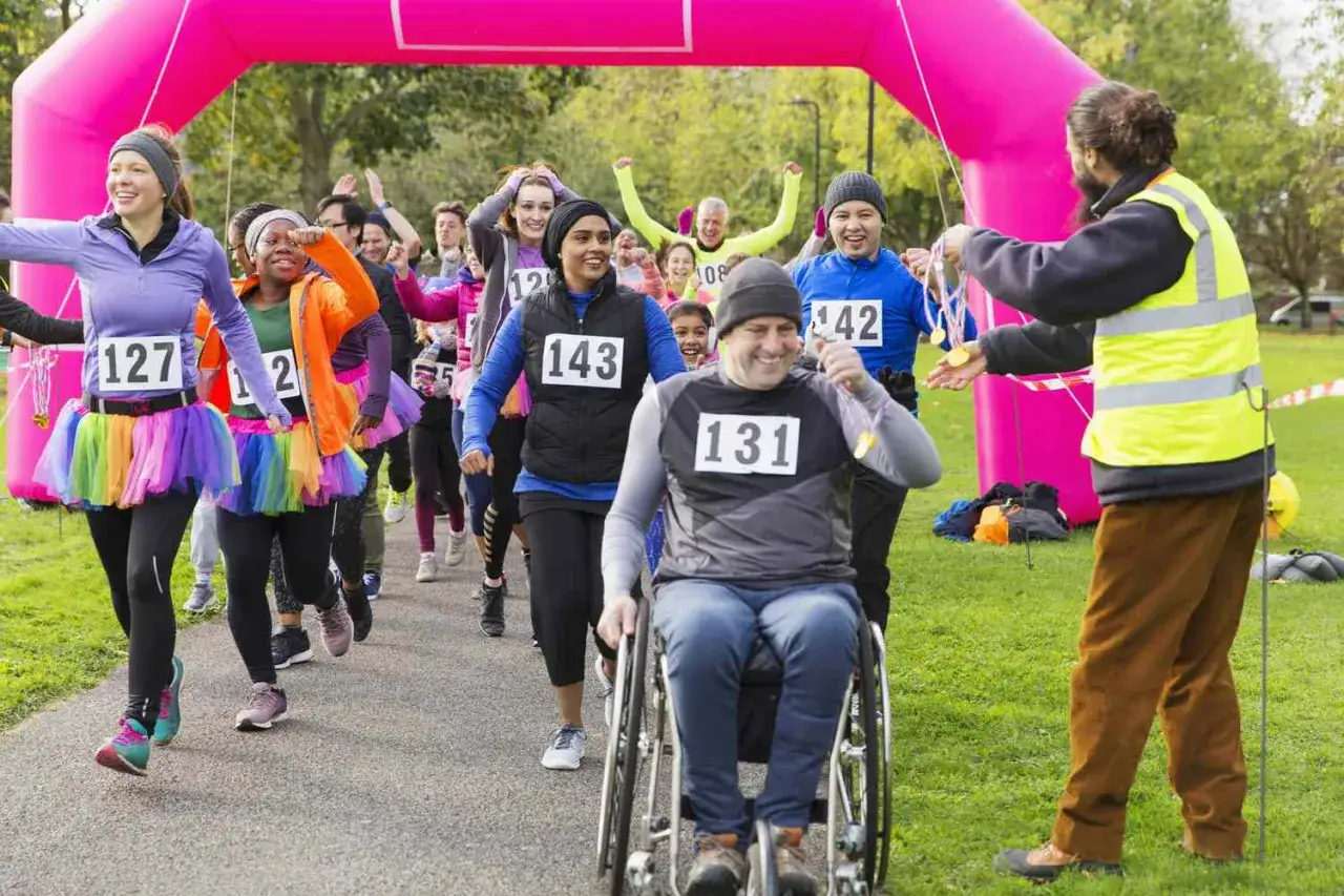Runners in colorful tutus and bibs cross a finish line. A man in a wheelchair smiles, showing easy fundraising ideas for individuals.