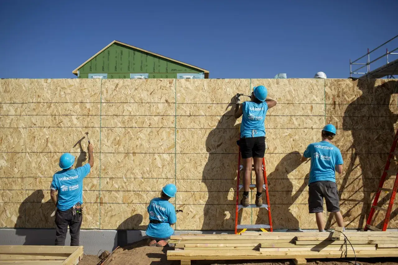 Volunteers in blue shirts and hard hats work on a house frame, embodying what is a service trip by building homes for Habitat for Humanity.