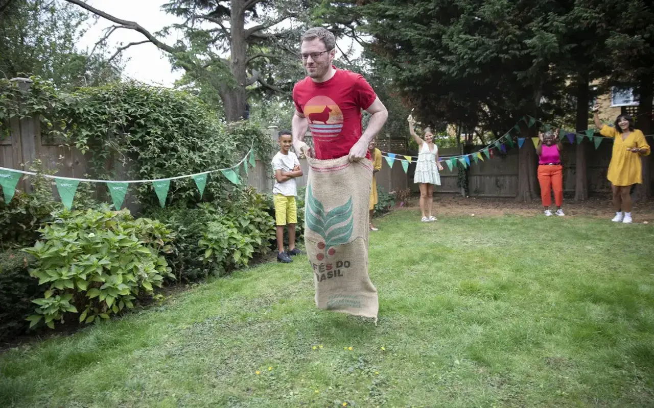 A man in a red shirt plays a sack race in a backyard, a fun idea for fundraising for 14 year olds. Other people watch and cheer.