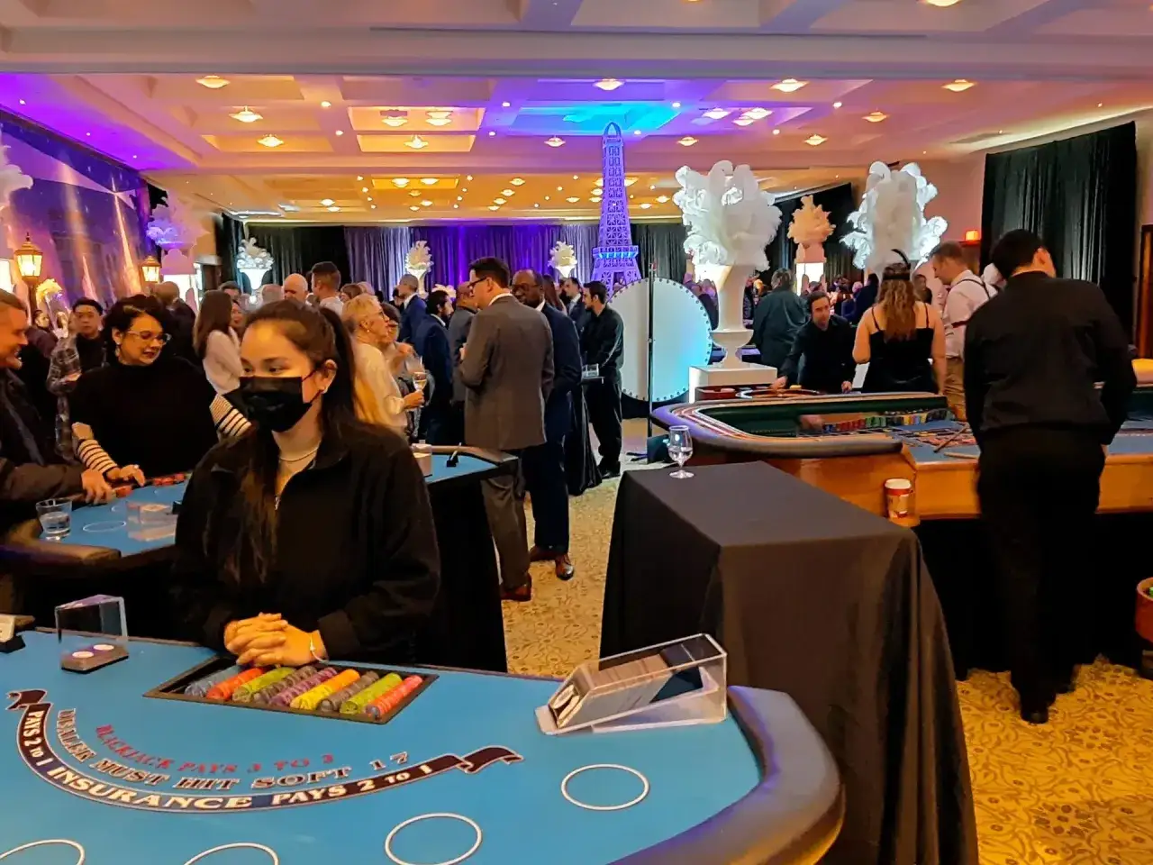 People enjoy a casino fundraiser with blackjack tables and a Paris-themed backdrop.
