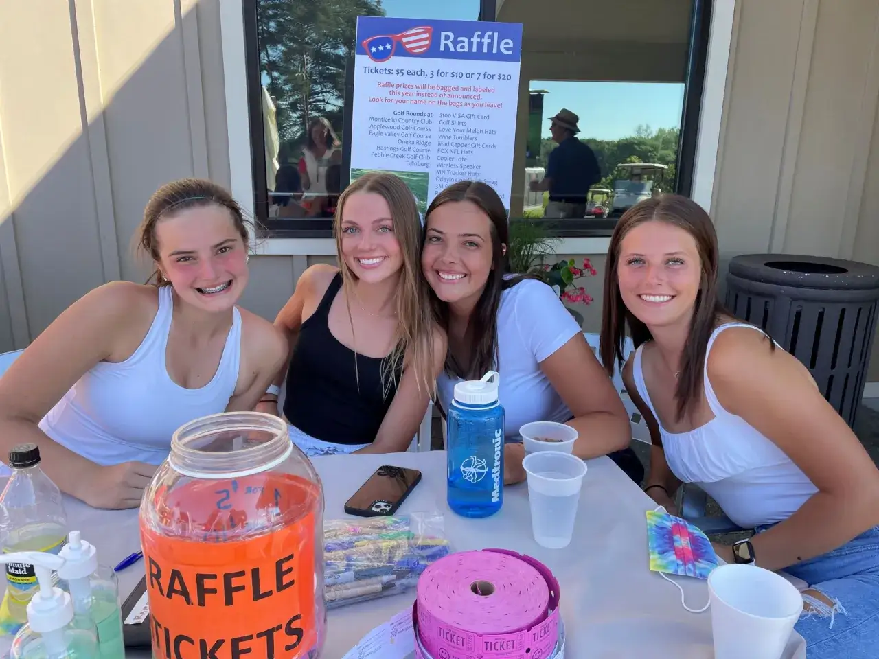Four smiling young women are at a table, ready to sell raffle tickets. A large jar labeled 