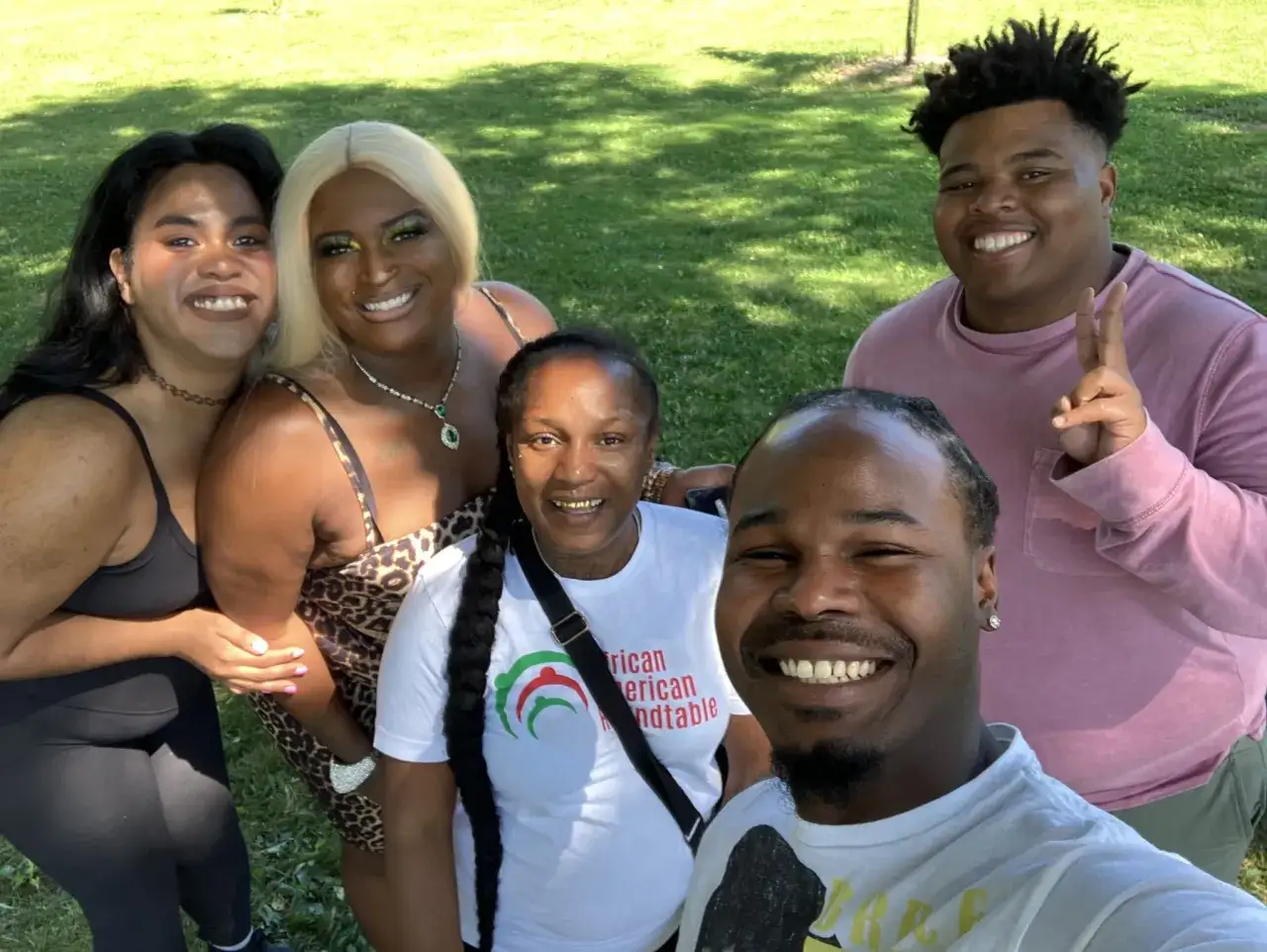 Five smiling people pose for a selfie outdoors. One woman wears a shirt that says