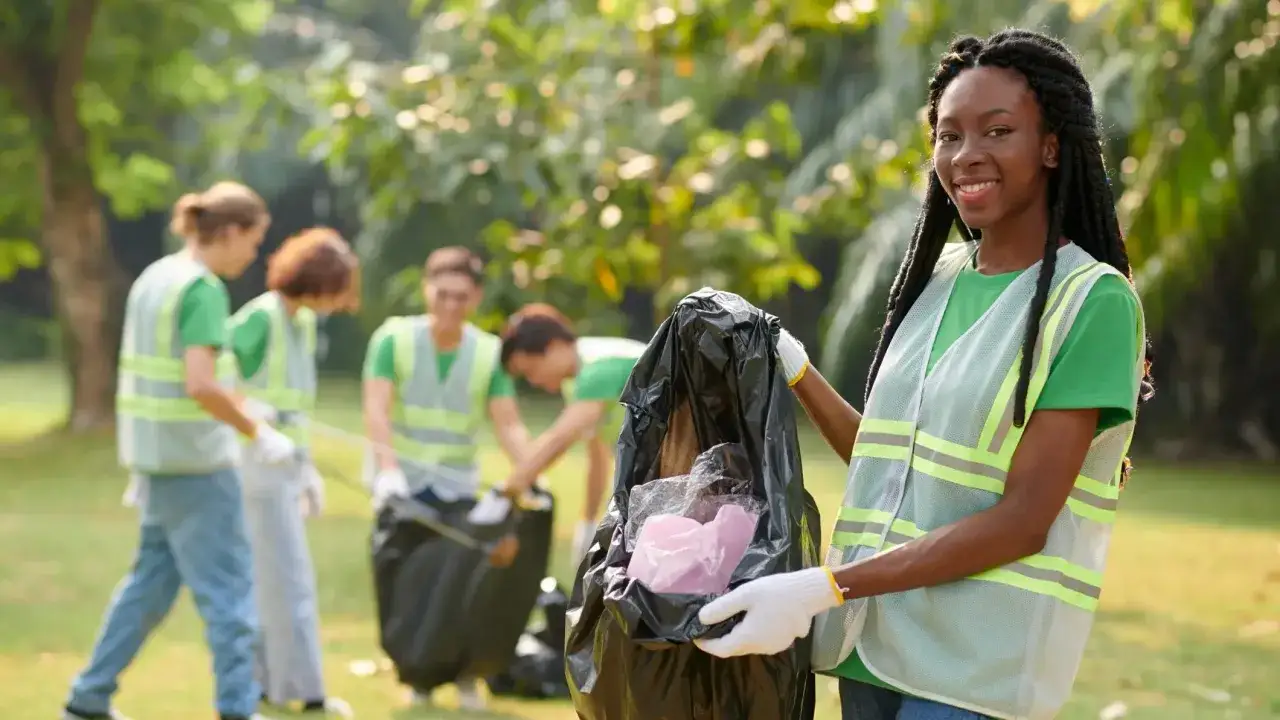 A smiling woman proudly holds a full trash bag, participating in voluntary work with her team cleaning a park.