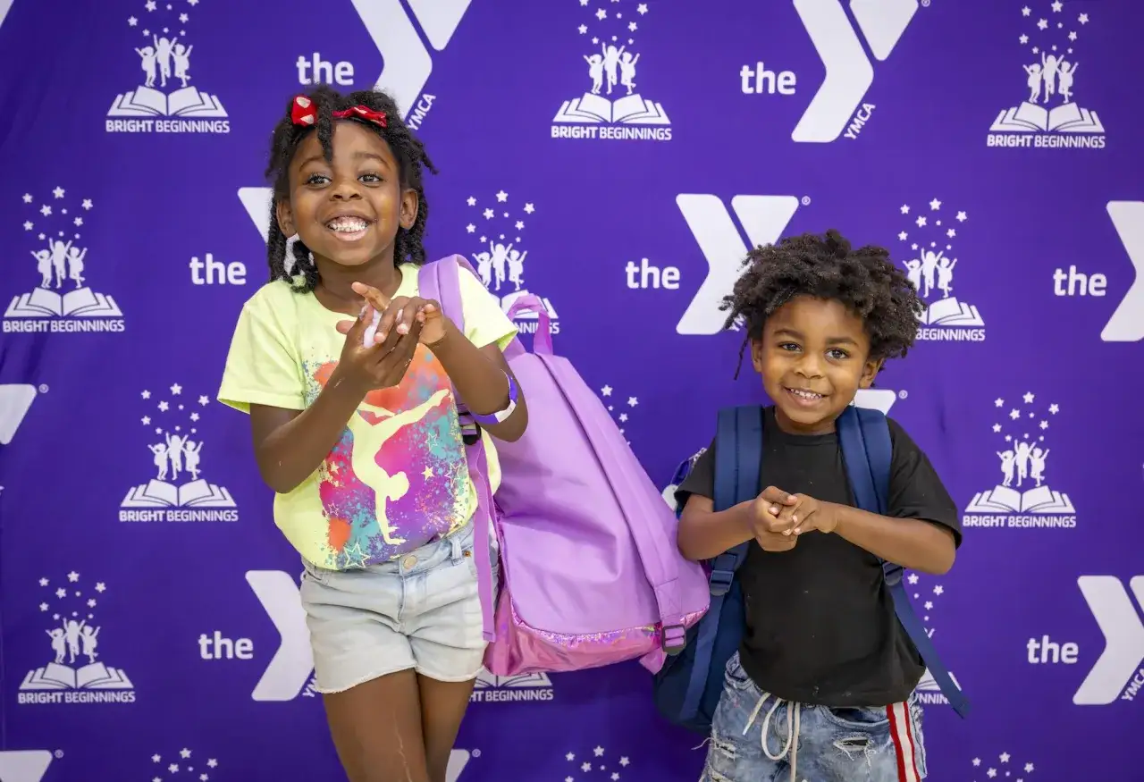 Two smiling children with backpacks stand in front of a purple backdrop with YMCA branding, ready for a YMCA fundraiser event.