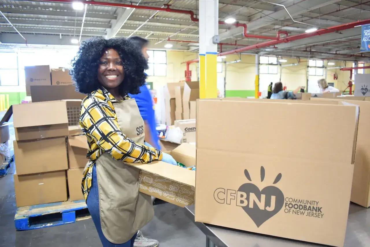 A smiling volunteer at the Community Foodbank of New Jersey, packing boxes. This image captures the essence of a food bank volunteer job description.