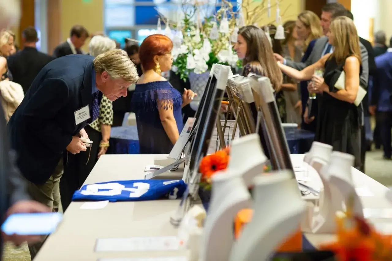 Guests examine items at a silent auction. A man in a suit leans over a table displaying a blue jersey and framed art.