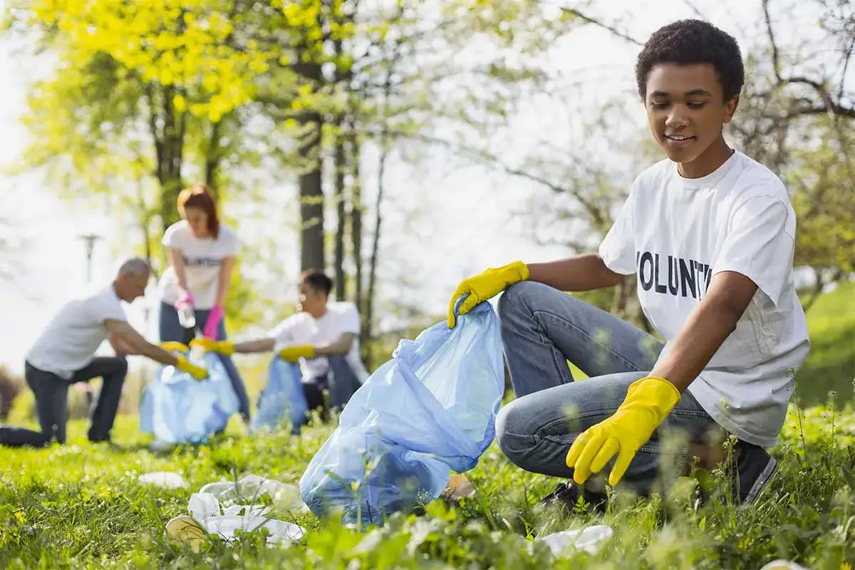 Young volunteers participate in a community project, cleaning up a park. They wear yellow gloves and collect litter in blue bags.
